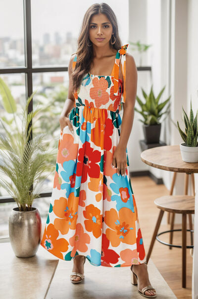 A woman wearing a floral print smocked dress with a square neckline, posing indoors with plants and furniture in the background.